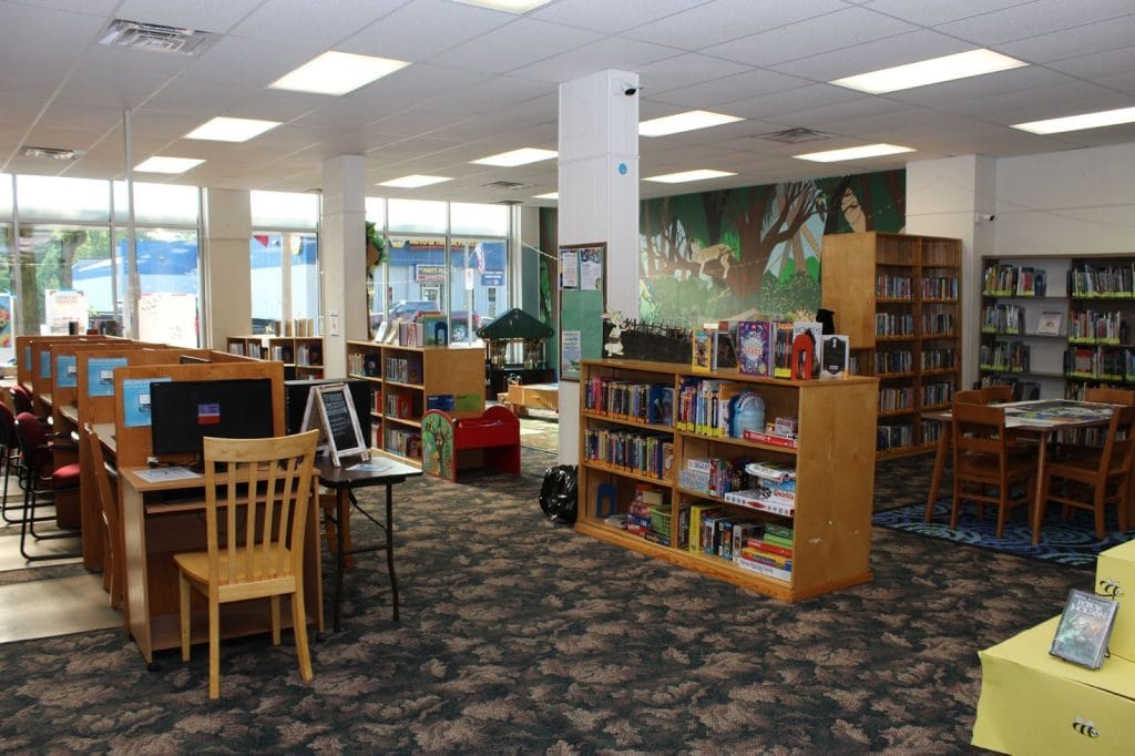 Interior of a library with bookshelves filled with books, a seating area with red chairs and a wooden table, and computer stations. The room is well-lit with large windows and has a colorful mural on one wall.