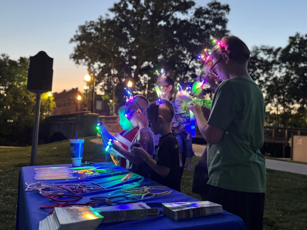 Children wearing glowing LED headbands examine toys and brochures on a table at dusk in a park. Trees and streetlights are visible in the background, creating a lively, festive atmosphere.
