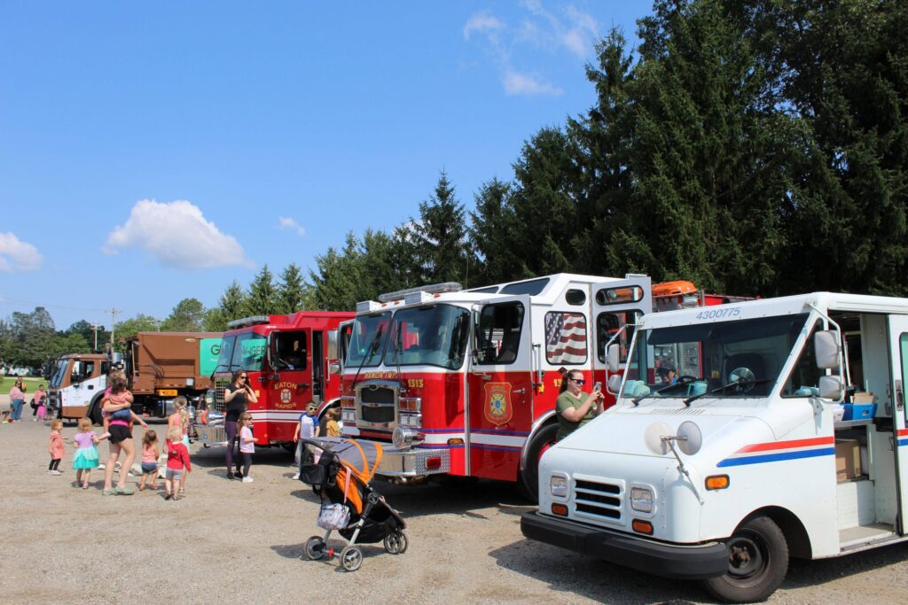 A group of people, including children, gather outdoors near fire trucks, a garbage truck, and a postal truck parked in a row on a sunny day with trees in the background.