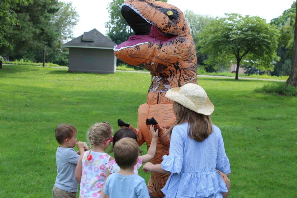 A group of young children and an adult wearing a sunhat gather around a person in an inflatable dinosaur costume in a grassy park with trees and a small building in the background.