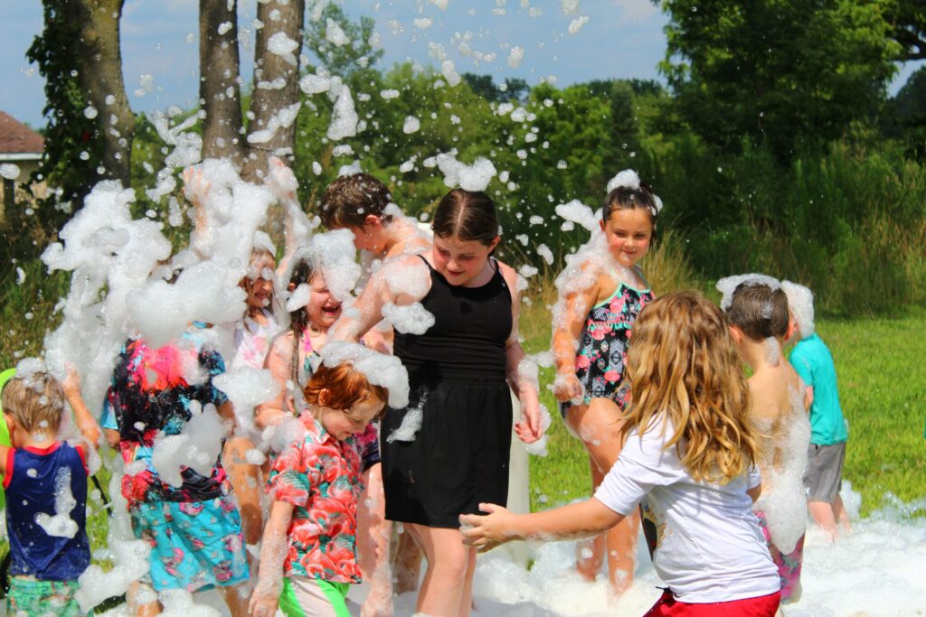 A group of children play in an outdoor area covered in foam, laughing and smiling on a sunny day. Some wear swimwear and colorful clothes, and green trees are visible in the background.
