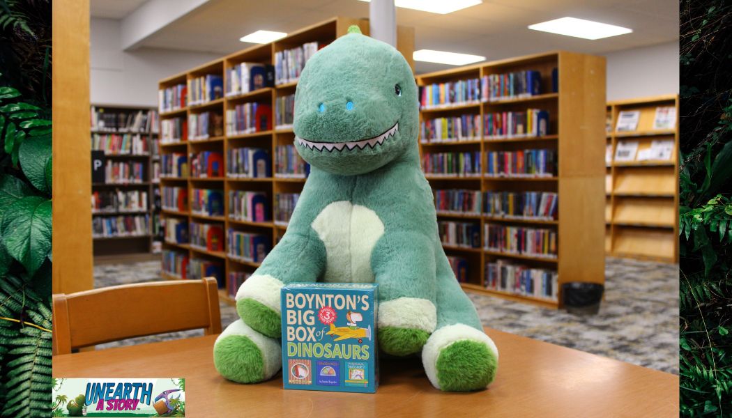 A large green plush dinosaur sits at a library table with a box set titled Boyntons Big Box of Dinosaurs in front of it. Bookshelves filled with books are visible in the background.