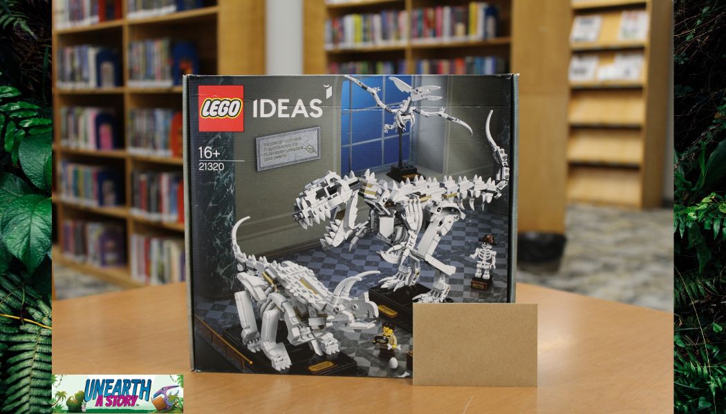 A LEGO Ideas set (21320) featuring dinosaur skeleton models sits on a wooden table in a library, with bookshelves in the background. A small brown card and the Unearth A Vision logo are visible in the image corners.