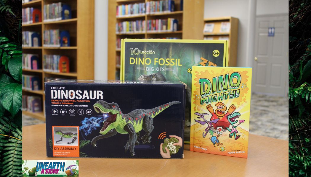 A photo of a dinosaur-themed DIY toy kit, a Dino Fossil dig kit, and the book Dino Mighty! displayed on a table in a library with bookshelves in the background.