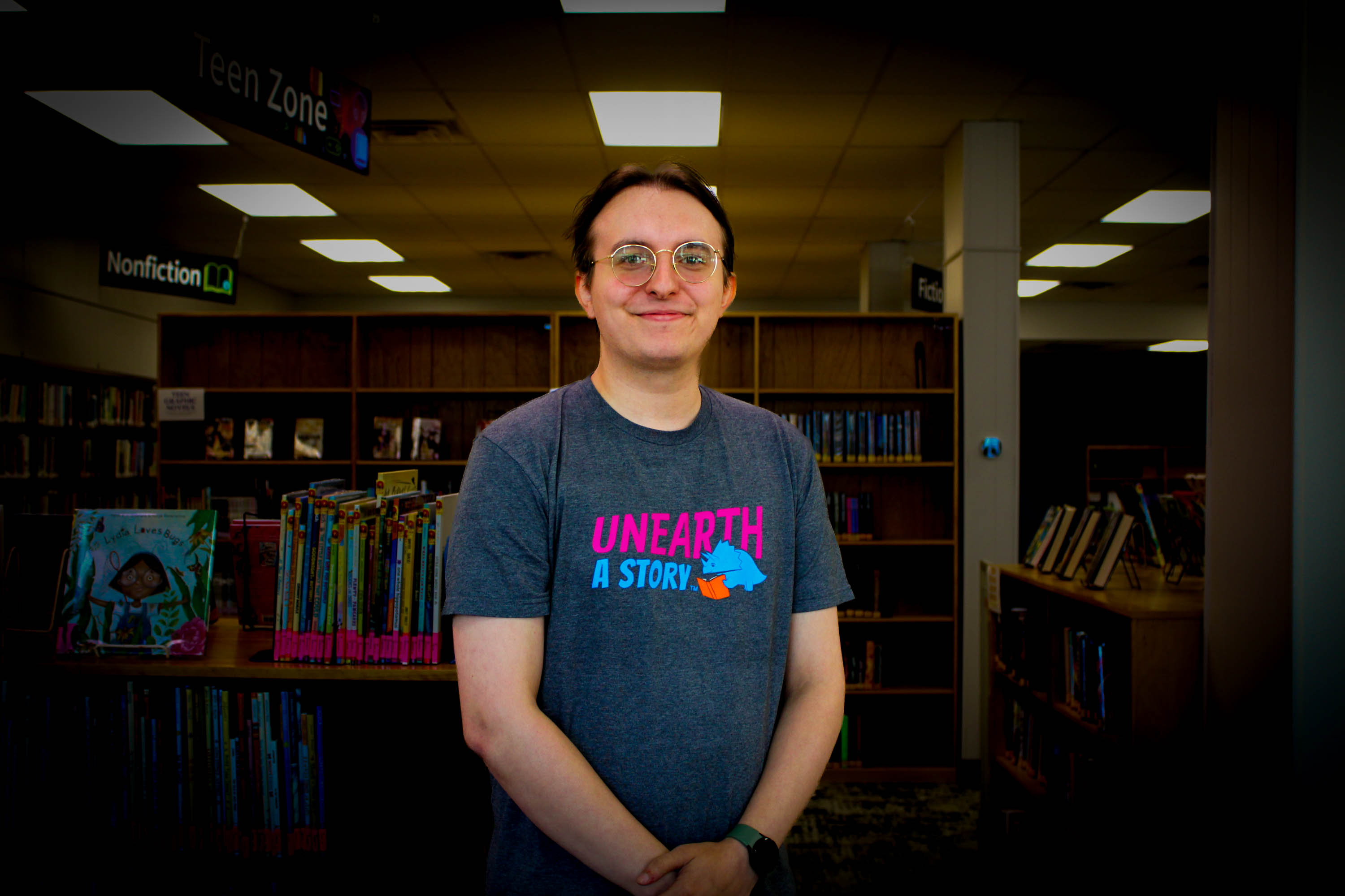 A person wearing glasses and a gray UNEARTH A STORY t-shirt stands smiling in a library, with bookshelves and a Teen Zone sign visible in the background.
